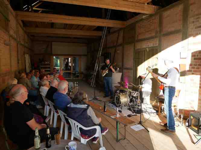 Band spielt, Publikum auf Bänken und Stühlen im Halbkreis davor.